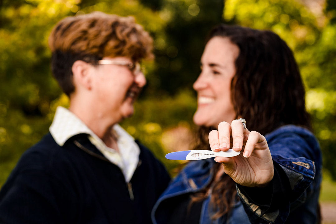 Wives hold up positive pregnancy test and smile at each other for baby announcement.